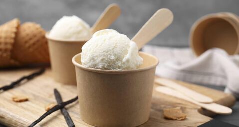 Paper cups with delicious ice cream and vanilla pods on wooden board, closeup